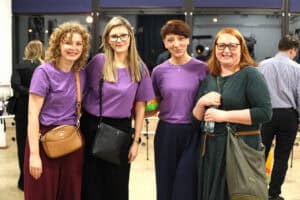 Cztery kobiety stoją obok siebie w sali. Trzy z nich mają na sobie fioletowe koszulki, a czwarta ciemnozieloną sukienkę. Kobiety pozują do wspólnego zdjęcia. Wszystkie są uśmiechnięte, patrzą wprost w obiektyw.
