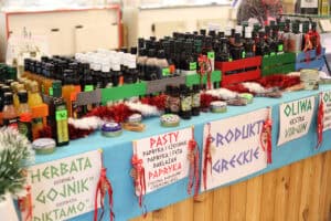 Stoisko z produktami greckimi przykryte błękitnym obrusem, udekorowane czerwono-białym łańcuchem choinkowym. Na blacie w kolorowych skrzynkach ustawiono liczne butelki z oliwą, octem i syropami. Z przodu lady wiszą białe tabliczki z napisami: „Herbata Górska Gojnik”, „Pasty: papryka i czosnek, bakłażan”, „Produkty greckie” oraz „Oliwa Extra Virgin”.