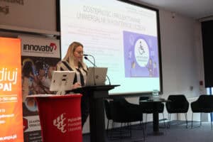 Prelegentka w pasiastej koszuli (Karolina Kowalczyk) mówi do mikrofonu przy mównicy. W tle na ekranie wyświetlany jest slajd: „Dostępność i projektowanie uniwersalne w kontekście uczelni”. Po prawej stronie slajdu widnieje fioletowa grafika z ikoną osoby na wózku inwalidzkim wewnątrz okręgu oraz napis „Projektowanie Uniwersalne”.