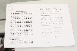 Zbliżenie na kartkę papieru, która jest materiałem szkoleniowym. Lewa strona zawiera schemat PISMA BRAILLE'A, w tym litery alfabetu, polskie znaki diakrytyczne oraz cyfry. Prawa strona zawiera ćwiczenia, w których należy uzupełnić luki w nazwach owoców, warzyw i miast, używając symboli Braille'a
