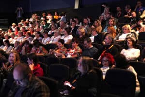 Szerokie ujęcie publiczności siedzącej w zaciemnionej kinowej sali, na ciemnych fotelach. Ludzie w różnym wieku, ubrani w stroje codzienne i biznesowe. Wśród publiczności znajdują się osoby patrzące na scenę, a także kilka osób korzystających z telefonów