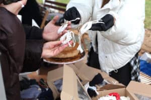 Zbliżenie na prezentację ozdób choinkowych. Osoba po prawej, ubrana w białą, puchową kurtkę i czarne rękawiczki, prezentuje małe, białe ozdoby świąteczne wykonane z plecionej, grubej włóczki, które trzyma w dłoniach. Druga osoba wyciąga dłoń w brązowym rękawie, aby obejrzeć lub odebrać rękodzieło. Na stole, w centrum kadru, znajduje się okrągłe ciasto z chrupiącą, brązową wierzchnią warstwą.