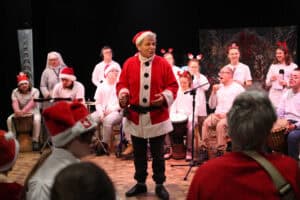 Występ muzyczny grupy Rozśpiewany Zakątek z Puław. Na drewnianej scenie gra i śpiewa duża grupa dorosłych i młodzieży w jasnych strojach i świątecznych nakryciach głowy; w centrum Michał Stanowski przebrany za Mikołaja. W tle widoczna jest zimowa dekoracja, a na pierwszym planie zarys widowni.