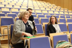 Kobieta w szarym kardiganie siedzi w auli wykładowej i trzyma mikrofon, drugą dłoń opiera na zielonym folderze leżącym na pulpicie. Obok niej siedzi druga kobieta w czarno-szarej bluzce, a za nimi widoczny jest mężczyzna w okularach. Tło stanowią puste rzędy niebieskich foteli z drewnianymi pulpitami.