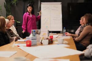 Prowadząca w fuksjowej koszuli wskazuje zielonym mazakiem na flipchart podzielony na cztery sekcje: „moje talenty”, „moje wartości”, „moje osiągnięcia”, „moje wsparcie”. W tle uczestniczki przy stole, na blacie kubki, arkusze, świeczniki i pudełka z chusteczkami.