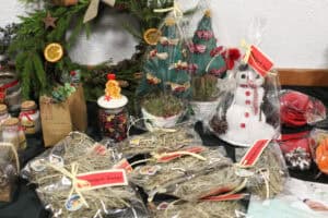 Zbliżenie na stół z upominkami świątecznymi: materiałowe choinki, bałwanek, słoiczki z „Zimowym Odprężeniem” (sole do kąpieli) oraz woreczki z siankiem z napisem „Wesołych Świąt”. W tle duży wieniec z suszonymi pomarańczami.