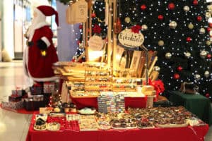 Stoisko z rękodziełem „DrewnoLuby” na tle dużej, oświetlonej choinki. Na pierwszym planie, na czerwonym obrusie, wyeksponowano bogaty wybór biżuterii: drewniane kolczyki na stojakach oraz liczne bransoletki z koralików i kamieni ułożone w rzędach. W tle widać drewniane deski z wypalanymi wzorami oraz dużą figurę Świętego Mikołaja z lampionem.