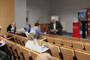 Moment wręczania dokumentów lub certyfikatów. Mężczyzna w brązowej marynarce (Wojciech Dec) pochyla się w stronę uczestniczki siedzącej w drugim rzędzie, podaje jej kartkę lub dyplom i wymienia uścisk dłoni. W tle widoczna jest czerwona mównica z logo WSPA oraz banery informacyjne.