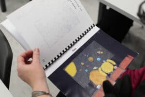 Zbliżenie na dłonie trzymające otwartą książkę (prawdopodobnie dla dzieci lub edukacyjną) w formacie Braille'a. Lewa strona książki zawiera tekst z wypukłymi punktami w alfabecie Braille'a. Prawa strona przedstawia ilustrację zatytułowaną "Kino" – ciemną scenę kosmosu z planetami i rakietami, z dodatkową siatką dotykową i materiałami o różnej teksturze