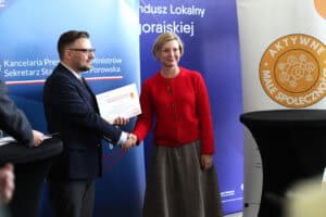 Ceremonia wręczenia certyfikatów. Mężczyzna (Piotr Stec) w granatowej marynarce i okularach ściska dłoń kobiety w czerwonym kardiganie i brązowej, długiej spódnicy, wręcza jej certyfikat. Tło stanowią banery informacyjne z napisami takimi jak "Fundusz Lokalny" i logo "Aktywne Małe Społeczności"