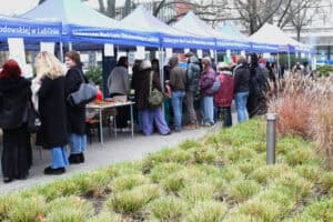 Szerokie ujęcie plenerowe długiego szeregu niebieskich namiotów z napisem Uniwersytet Marii Curie-Skłodowskiej w Lublinie, ustawionych na chodniku. Przed namiotami, wzdłuż stołów stoisk kiermaszu, ciągnie się długa kolejka ludzi w ciepłych, zimowych ubraniach. W dolnej części kadru widoczny jest pas gęstej, jasnozielonej trawy ozdobnej.