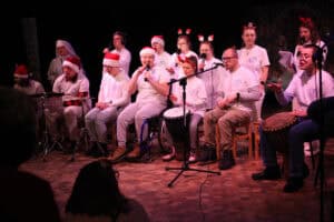 Występ muzyczny grupy Rozśpiewany Zakątek z Puław. Około 14 osób, głównie młodzież i dzieci wraz z zakonnicą, siedzi w półkolu na scenie i śpiewa; w centrum mężczyzna na wózku trzyma mikrofon, a część uczestników gra na instrumentach perkusyjnych. W tle widoczne są dyskretne dekoracje świąteczne.