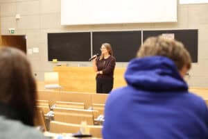 dr inż. Agnieszka Gandzel stoi przodem do słuchaczy w auli i przemawia do mikrofonu. Nad kobietą widać dolną część białego ekranu projekcyjnego oraz fragment drewnianego biurka. Widok z perspektywy uczestnika.