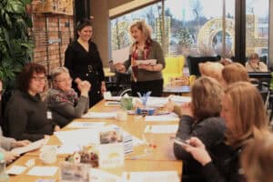 Wokół długiego stołu siedzą uczestniczki. W tle dwie prowadzące: w czarnej sukience i w oliwkowej bluzce z bordową apaszką, trzymają białe dyplomy. Za szybą widać zimowy krajobraz i dekoracje świąteczne. Na stole materiały warsztatowe i kubki, po lewej zielone rośliny.