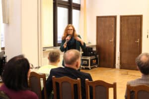 Kobieta (Marta Chruściel) o długich, falowanych włosach i w okularach, ubrana w ciemną marynarkę, stoi przed publicznością, trzyma mikrofon i przemawia. Publiczność, widoczna od tyłu, siedzi na drewnianych krzesłach. Za kobietą, w tle, widoczne są duże okna; ciemne, drewniane drzwi oraz czarne skrzynie ze sprzętem technicznym na małym wózku z logo "TLN"