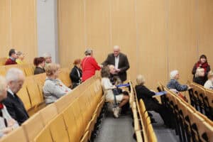 Wojciech Dec stoi w przejściu między rzędami drewnianych krzeseł. Pochyla się w stronę kobiety w czerwonym swetrze, wręczając jej biały dokument (certyfikat). Kobieta wyciąga rękę, by go odebrać. W rzędach obok siedzą inni uczestnicy; niektórzy z uwagą obserwują tę scenę, inni rozmawiają. Po prawej stronie kadru, w tle przy ścianie, stoi kobieta w ciemnym stroju z czerwonym szalikiem, trzymająca mikrofon i obserwująca sytuację. Tło stanowią jasne, drewniane panele ścienne.