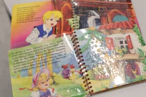 Rozkładówka starej książki w oprawie na spiralce. Strona po lewej przedstawia kolorowe ilustracje i tekst, w tym fragmenty z bajek o trzech misiach oraz o blond chłopcu otwierającym drzwi szafy. Obie strony są pokryte folią transparentną z wypukłościami