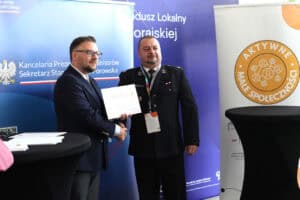 Ceremonia wręczenia certyfikatów. Mężczyzna (Piotr Stec) w granatowej marynarce i okularach ściska dłoń drugiego mężczyzny ubranego w czarny galowy mundur (strażacki), wręcza mu certyfikat. Tło stanowią banery informacyjne z logo Kancelarii Prezesa Rady Ministrów i okrągłym logo "Aktywne Małe Społeczności"
