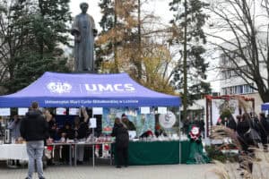 Szerokie ujęcie plenerowe dużego, niebieskiego namiotu z białym napisem „UMCS Uniwersytet Marii Curie-Skłodowskiej w Lublinie". Namiot ustawiony jest pod wysokim, szarym pomnikiem Marii Curie-Skłodowskiej, stojącym na postumencie. Przy stołach pod namiotem stoi grupa ludzi. Po prawej stronie stoiska, na dole, stoi duża, pluszowa figura Świętego Mikołaja w czerwonym płaszczu i zielonym worku.