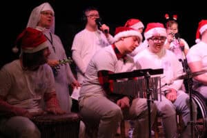 Świąteczny występ muzyczny na scenie. Grupa dorosłych i młodzieży w białych koszulkach i czapkach Mikołaja gra na instrumentach perkusyjnych; w centrum siedzi młody mężczyzna pochylony nad instrumentem. Po lewej stoi zakonnica z grzechotką, a scena jest oświetlona ciepłym światłem.