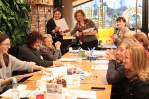 Uczestniczki siedzą przy długim stole. W głębi dwie prowadzące przekazują arkusz papieru. Po prawej kobieta bije brawo. Na stole materiały i kubki. W tle przez szybę widać zimowe dekoracje świąteczne.