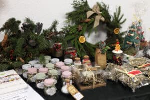 Ekspozycja różnorodnych prezentów na stole: słoiczki z ziołami i herbatami z kolorowymi zakrętkami, stroiki z igliwia oraz książki. W centralnej części widoczne jest pudełko z zestawem zapachowym, udekorowane sznurkiem i suszonymi roślinami.