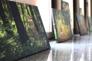 Ujęcie w perspektywie zbieżnej. Zbliżenie na serię dużych, prostokątnych wydruków zdjęć na płótnie, ustawionych rzędem na błyszczącym, nakrapianym parapecie. Pierwszy plan skupia się na zdjęciu przedstawiającym kobietę w lesie, ubraną w długą suknię wykonaną z naturalnych elementów, takich jak mech lub liście. Zdjęcia w tle są nieostre, a za nimi widoczne są pionowe ramy okienne, wpuszczające jasne światło