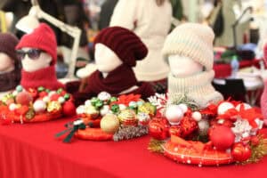 Na czerwonym obrusie wyeksponowano trzy dekoracyjne stroiki w formie wianków, bogato zdobione kolorowymi bombkami (czerwonymi, złotymi i srebrnymi) oraz sztucznymi kwiatami gwiazdy betlejemskiej. W tle, na białych styropianowych głowach ekspozycyjnych, zaprezentowano ręcznie robione, wełniane czapki i kominy w kolorach czerwonym, bordowym oraz białym.