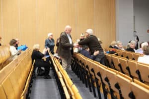 Moment gratulacji podczas wydarzenia. W centrum kadru mężczyzna w brązowej marynarce (Wojciech Dec) wymienia uścisk dłoni z jednym z uczestników – starszym mężczyzną, który wstał ze swojego miejsca w rzędzie. Obaj mężczyźni są zwróceni ku sobie profilami. Wokół nich widoczni są inni uczestnicy konferencji.