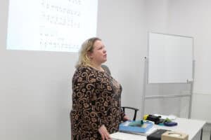 Kobieta (Ewelina Orzechowska) w czarno-złotej, wzorzystej bluzce stoi obok biurka, przemawia. W tle za nią wyświetla się schemat pisma Braille'a, a po prawej stronie stoi pusta, biała tablica suchościeralna. Na biurku leżą materiały szkoleniowe, w tym niebieskie pudełko
