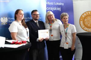 Zdjęcie grupowe z ceremonii wręczenia nagród. Mężczyzna (Piotr Stec) w granatowej marynarce trzyma certyfikat, pozując do zdjęcia w otoczeniu trzech kobiet w białych koszulkach i czarnych spódnicach/spodniach, które mają przypięte identyfikatory, a na ich szyjach znajdują się kolorowe smycze. W tle widoczne są banery z logotypami Kancelarii Prezesa Rady Ministrów oraz „Aktywne Małe Społeczności"