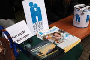 Stoisko poświęcone zbiórce charytatywnej na rzecz Lubelskiego Hospicjum dla Dzieci im. Małego Księcia. Na środku stołu stoi duży, biały plakat z logo Hospicjum. Obok niego leży mały stos broszur, w tym magazyn „Mały Książę" oraz książeczka „Pięć Lekcji z Krainy Marzeń" w ciemnej oprawie. Na małej, białej karteczce po lewej widnieje drukowany napis: „Pierniczki 5 zł/zestaw".