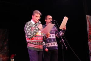 Dwóch dorosłych mężczyzn śpiewa na ciemnej scenie do mikrofonów, trzymają kartki z tekstem. Obaj mają świąteczne swetry z motywem reniferów; mężczyzna z lewej nosi okulary. W lewym dolnym rogu widać fragment świątecznej dekoracji w kształcie pingwina w czapce Mikołaja.