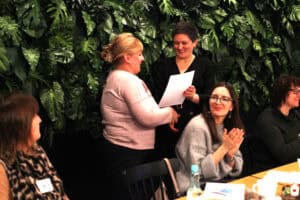 Wręczenie dokumentu na tle ściany z zielonymi liśćmi. Kobieta w czarnej sukience przekazuje biały arkusz drugiej kobiecie w szarym swetrze. Na pierwszym planie uczestniczki klaszczą.