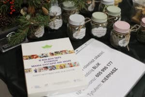 Zbliżenie na białe pudełko z napisem: „O rolach taty, których mama zastąpić nie może – karty motywacyjne dla ojców”. W tle słoiczki z siankiem i owocami i suszonymi ziołami, z etykietami takimi jak „Dobre relacje...”.