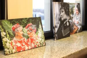 Seria portretów na płótnie ustawionych na nakrapianym, granitowym parapecie przy oknie. Na pierwszym planie znajduje się kolorowy portret dwóch starszych kobiet uśmiechających się do siebie, ubranych w czerwone bluzki z kwiecistym wzorem, naszyjniki z pereł i ekstrawaganckie nakrycia głowy, tzw. fascynatory. W tle widoczne są dwa kolejne portrety, przedstawiające kobiety w stylizowanych strojach