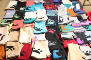 Kolekcja kolorowych, dzianinowych rękawiczek bez palców (mitenek) ułożonych w równych rzędach na czerwonym podłożu. Rękawiczki wykonane są z wełny w kolorach czarnym, beżowym, szarym oraz błękitnym i posiadają charakterystyczne, ręcznie haftowane wzory: kocie łapki, sylwetki kotów oraz kolorowe kwiaty.