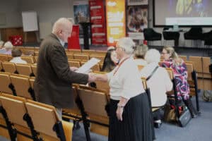 Moment wręczenia certyfikatu. Po lewej stronie stoi Wojciech Dec, który lewą ręką trzyma biały arkusz papieru, a prawą ściska dłoń starszej kobiety. Kobieta stoi przodem do niego, jest uśmiechnięta, ubrana w biały sweter i czarną, plisowaną spódnicę, a jej szyję zdobią czerwone korale. W tle widać fragmenty auli oraz grupę rozmawiających uczestniczek w dalszym planie. Na ścianie w głębi widoczne są materiały promocyjne uczelni, w tym duży pomarańczowy baner z napisem „Studiuj w WSPA”.
