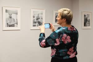 Kobieta o krótkich blond włosach i w bluzce w różowe i turkusowe kwiaty, stoi tyłem do widza i robi zdjęcie smartfonem jednej z czarno-białych fotografii wiszących w rzędzie na jasnej ścianie. W tle widoczne są cztery oprawione portrety