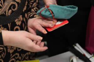 Zbliżenie na dłonie. Jedna osoba (w czarno-złotym wzorzystym stroju) trzyma stos czerwonych kart do gry. Druga osoba, widoczna fragmentarycznie, wręcza jej turkusowy woreczek ściągany pomarańczowym sznurkiem, leżący na kartach