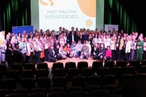 Zdjęcie grupowe uczestników i organizatorów konferencji „Moc Małych Społeczności" – stojących i siedzących na scenie w kilku rzędach, tzw. kaskadowo. W centralnym punkcie stoją mężczyźni w garniturach, a wokół nich osoby w różnym wieku. W tle widoczny jest ekran z hasłem konferencji. Na pierwszym planie widać puste rzędy czarnych foteli publiczności