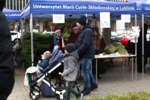 Rodzina przy stoisku pod niebieskim namiotem UMCS. Mężczyzna w czapce pcha wózek dziecięcy, obok niego idzie mały chłopiec. Za stołem, pod namiotem, stoją trzy uśmiechnięte młode kobiety, dwie z nich mają na głowach czerwone czapki Mikołaja, a jedna ciemną opaskę z rogami renifera.