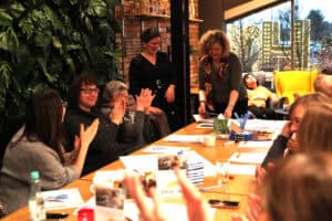 Po lewej stronie, na tle ściany z roślin, siedzą trzy uczestniczki i klaszczą. W głębi dwie prowadzące przy stole, pochylone nad dokumentami. W tle przez okno widoczny żółty fotel i świąteczne dekoracje. Na pierwszym planie rozmyte dłonie klaszczących osób.