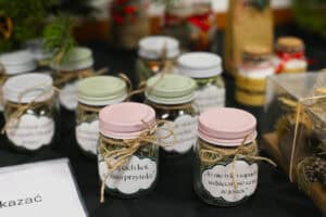 Zbliżenie na szklane słoiczki wypełnione suszonymi ziołami, ozdobione sznurkiem. Na etykietach widnieją napisy: „Ten zapach koi, a cisza przytula” oraz „To nie tylko zapach, wdzięczność za to, że jesteś”. Słoiczki mają kolorowe zakrętki: różowe i miętowe