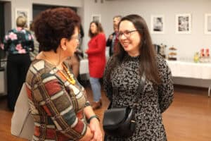 Zbliżenie na dwie uśmiechnięte kobiety rozmawiające podczas wernisażu. Kobieta po prawej, z długimi ciemnymi włosami i w okularach, nosi wzorzystą czarną sukienkę i czarną torebkę na ramieniu, patrzy na rozmówczynię. Druga kobieta (po lewej) ma krótkie, kręcone włosy i jest ubrana w kraciastą sukienkę z pomarańczowym naszyjnikiem. W tle widać stoły z poczęstunkiem i niewyraźne sylwetki innych gości