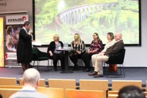 Grupa pięciu panelistów (Małgorzata Zmysłowska, Karolina Kowalczyk, Justyna Spryszak, Dorota Krać, Wojciech Dec) siedzi na scenie podczas konferencji. Po lewej stronie stoi kobieta w czarnej sukience (dr Marta Komorska), która przemawia do mikrofonu, trzymając w ręku kartkę. Pozostałe cztery osoby siedzą w fotelach wokół małego stolika z wodą mineralną. W tle widoczny jest duży ekran wyświetlający kolorowe zdjęcie portu z łodziami oraz baner z napisem „innovativ”.