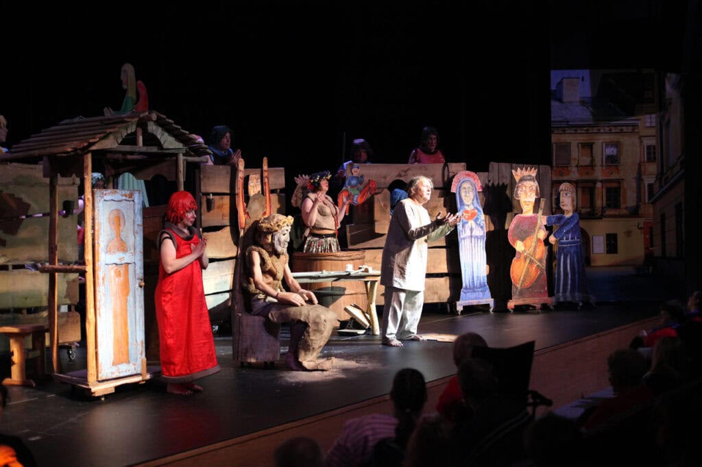 Punktowo oświetlona scena teatralna z elementami religijnymi i ludowymi. Po lewej stoi aktorka w czerwonej, długiej sukni z chustą na głowie, z rękami złożonymi w geście modlitwy, obok drewnianej budki z ikoną. W centrum, na pierwszym planie, siedzi na pniu aktor w brązowym kostiumie z maską. Po prawej stoi starszy mężczyzna w jasnej szacie, z rękami uniesionymi do góry, przemawia. W tle widać duże, płaskie, malowane postacie świętych oraz fragment miejskiej zabudowy.