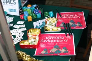 Na stole nakrytym zielonym obrusem leżą dwie czerwone ulotki z napisem „Akademicki Jarmark Świąteczny" oraz datą 4 grudnia 2025 r. Obok nich znajdują się czarne długopisy. Na środku stołu stoją dwie metalowe, czerwone formy w kształcie buta Mikołaja wypełnione kolorowymi cukierkami w żółtej i zielonej folii.