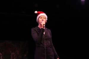 Występ muzyczny Klary Siuciak. Młoda kobieta w czapce świątecznej stoi na scenie i śpiewa do mikrofonu. Ma krótkie, jasne włosy, zamknięte oczy i ciemny strój. Scena jest oświetlona ciepłym światłem, a w lewym dolnym rogu widać fragment dekoracji świątecznej.