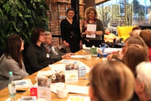 Uczestniczki siedzą przy stole. W centrum dwie prowadzące: jedna w czarnej sukience patrzy w stronę grupy, druga w zielonej bluzce trzyma biały arkusz. Po lewej ściana z zielenią, po prawej duże okno z fragmentem miejskich iluminacji.